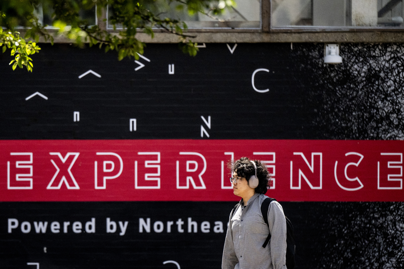 A person wearing headphones walks through campus with a wall in the background that says 'experience'.