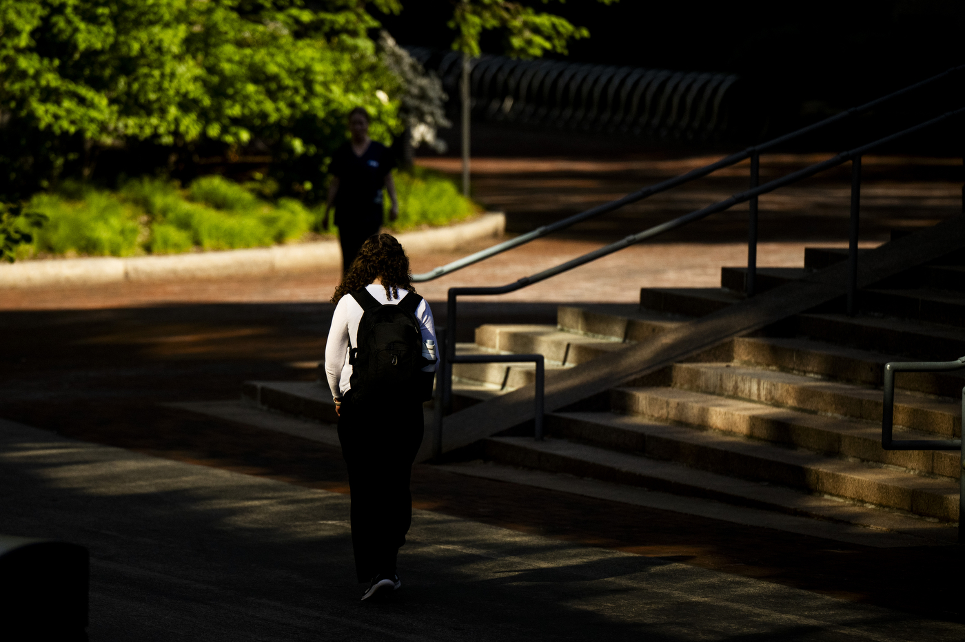 A student wearing a backpack walks through a shadowed part of campus.