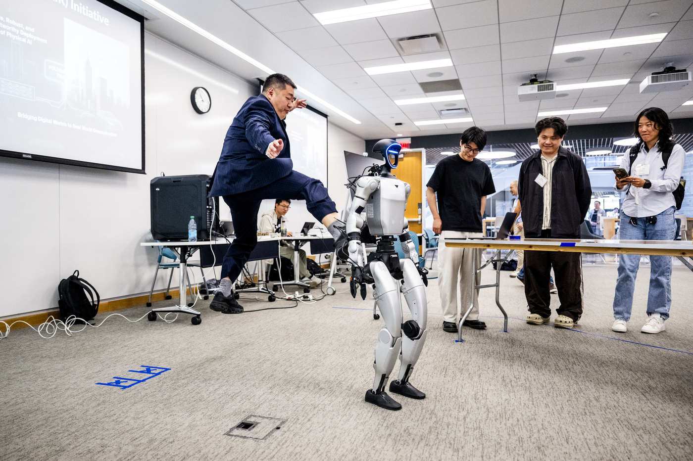 Students watch a presentation with a robot, featuring a person jumping in the air to kick the bipedal robot in the back.