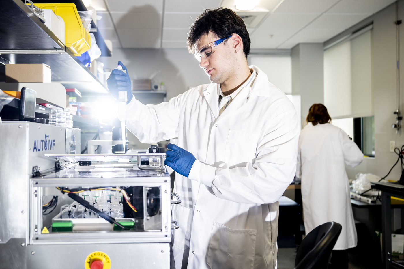A researcher in a white coat and safety glasses operating an AutoIVF machine in a lab.