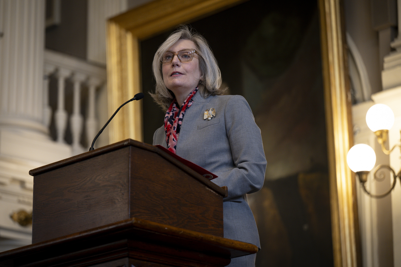 A low angle shot of a woman dressed in all gray addresses the crowd at a podium with a microphone.