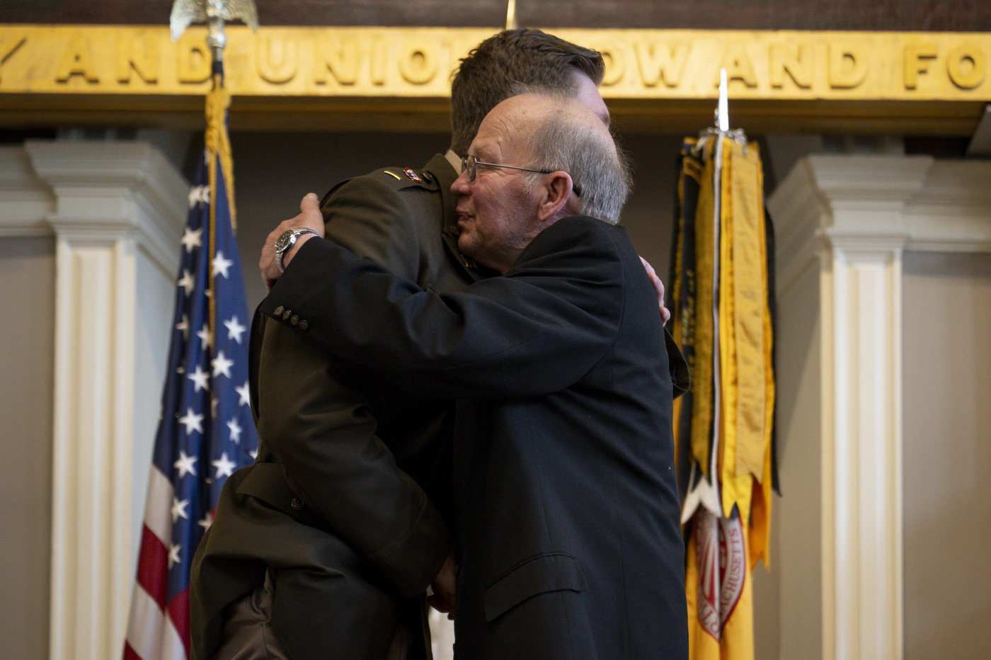 An older person embraces an ROTC cadet, with an American flag in the background.