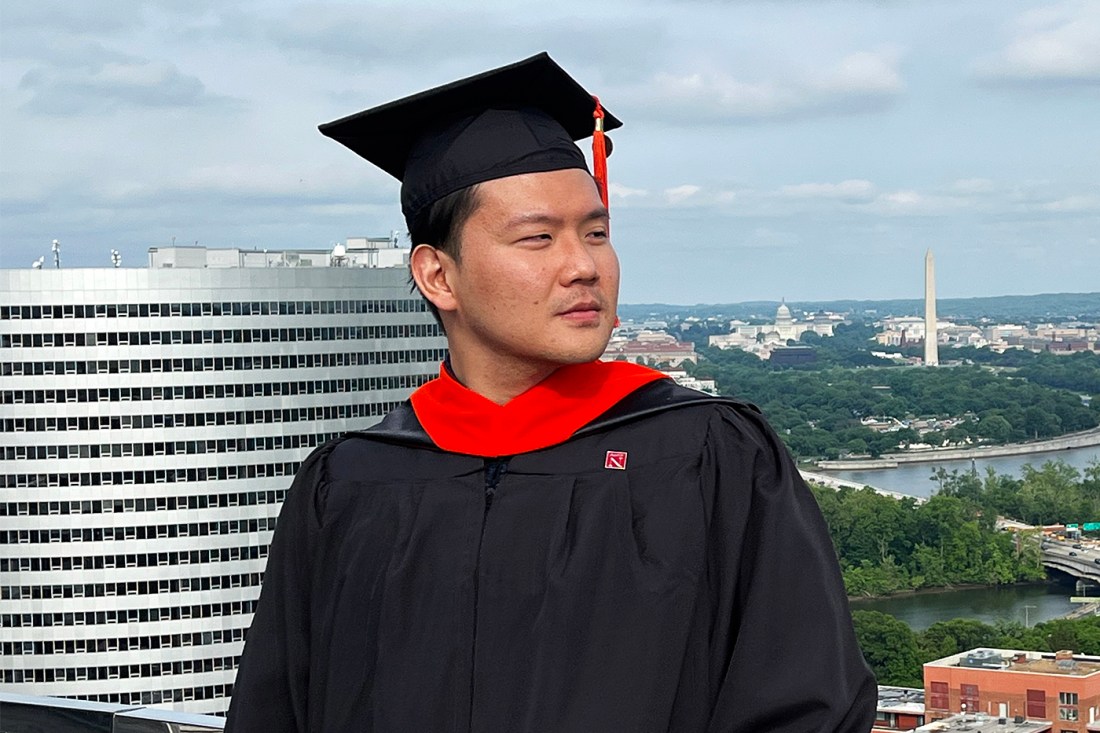 A man is wearing a cap and gown with a red hood and looking over his left shoulder while standing against the railing atop a tower.