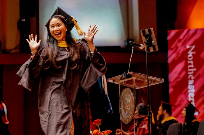 A happy graduate cross the stage at Northeastern Seattle's 2026 commencement while waving both hands.
