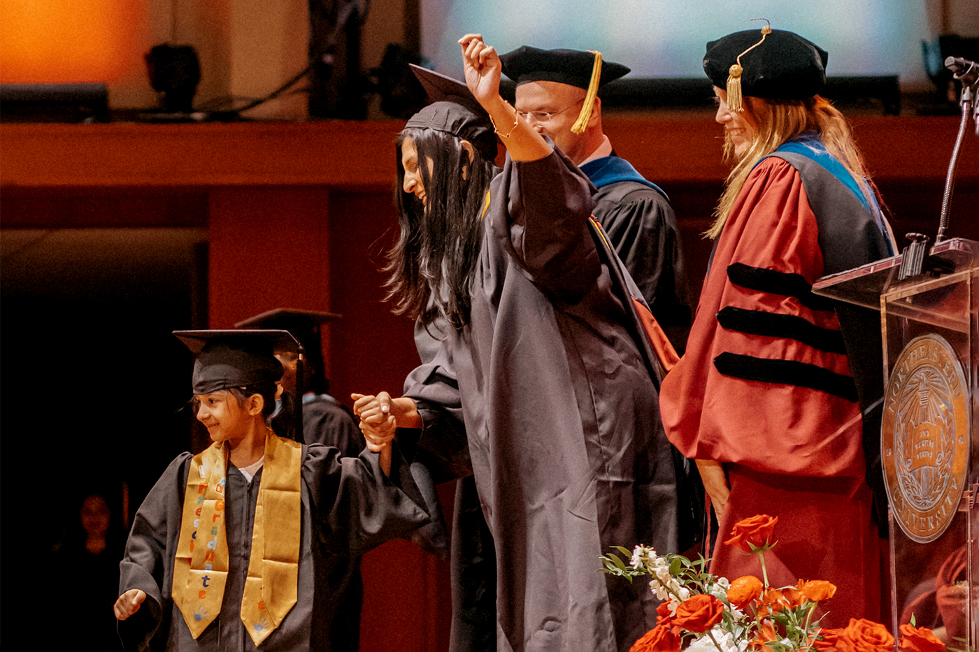A Northeastern graduate stands on stage alongside her child, both sporting caps and gowns.