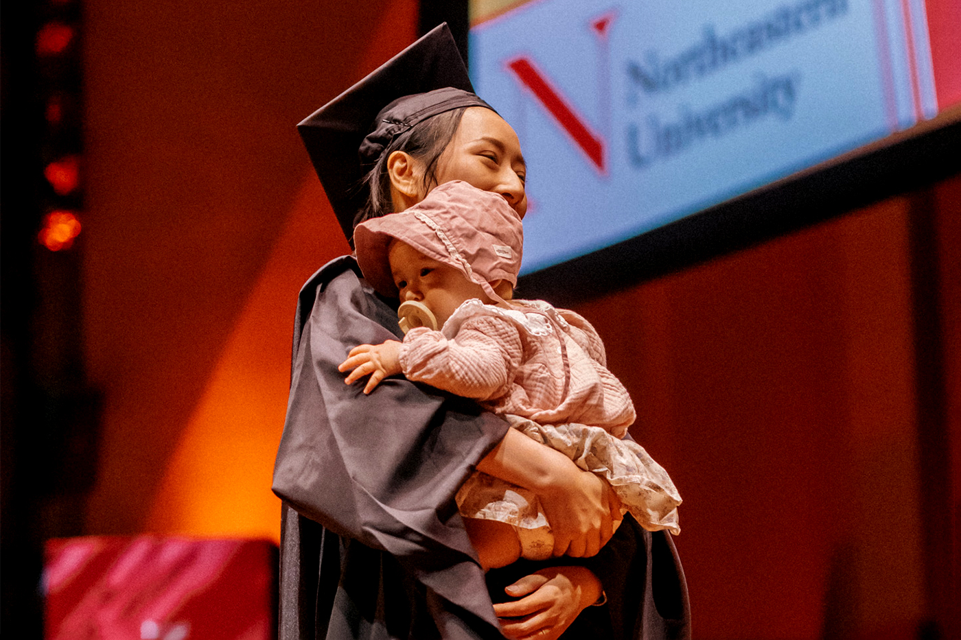 A Northeastern graduate wearing his cap and gown holds her baby. while