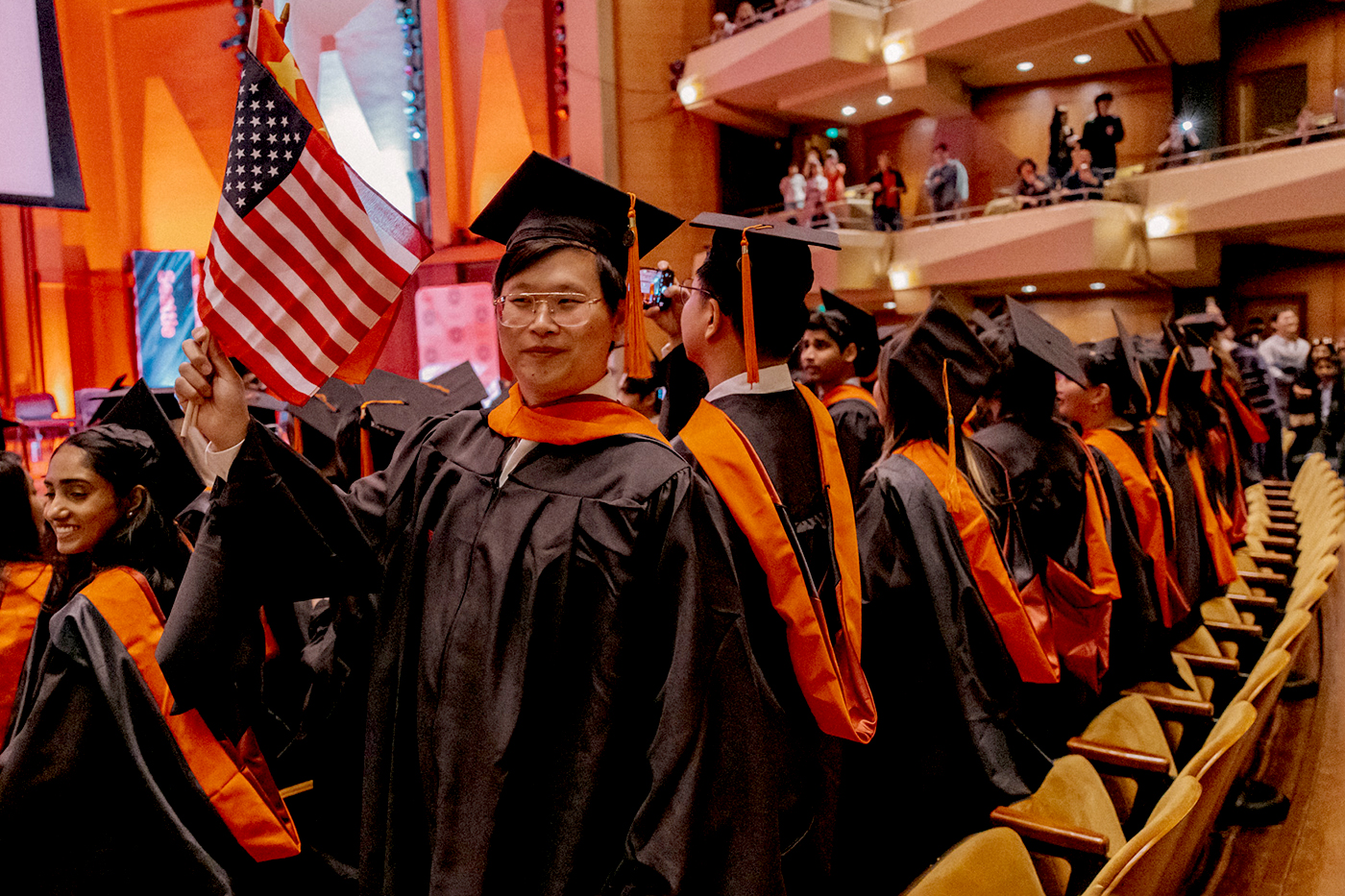 A group of Northeastern graduates, including one who waves an American flag, stand in front of their seats.