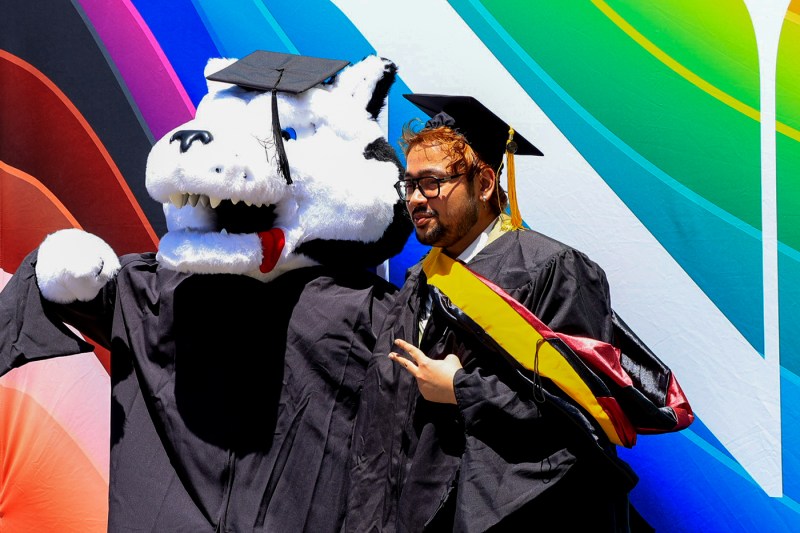 A graduate wearing regalia poses for a photo with Paws the mascot.