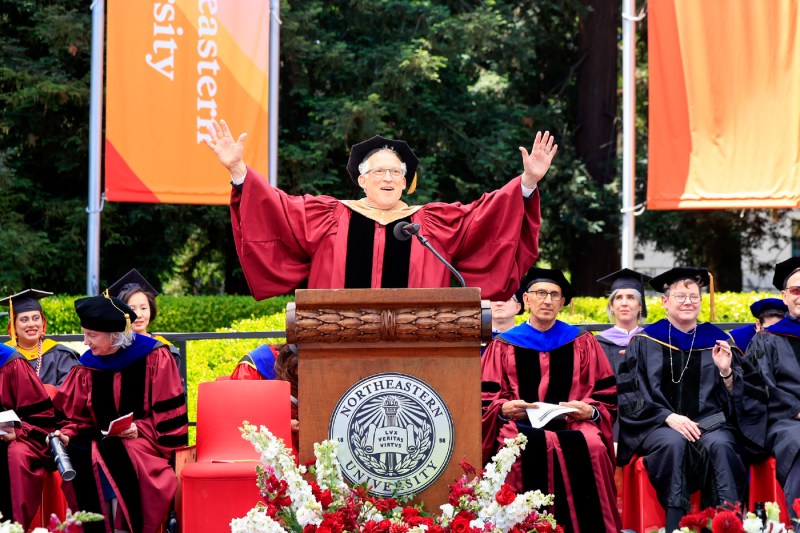 A faculty member in regalia stands at a podium with both arms outstretched.