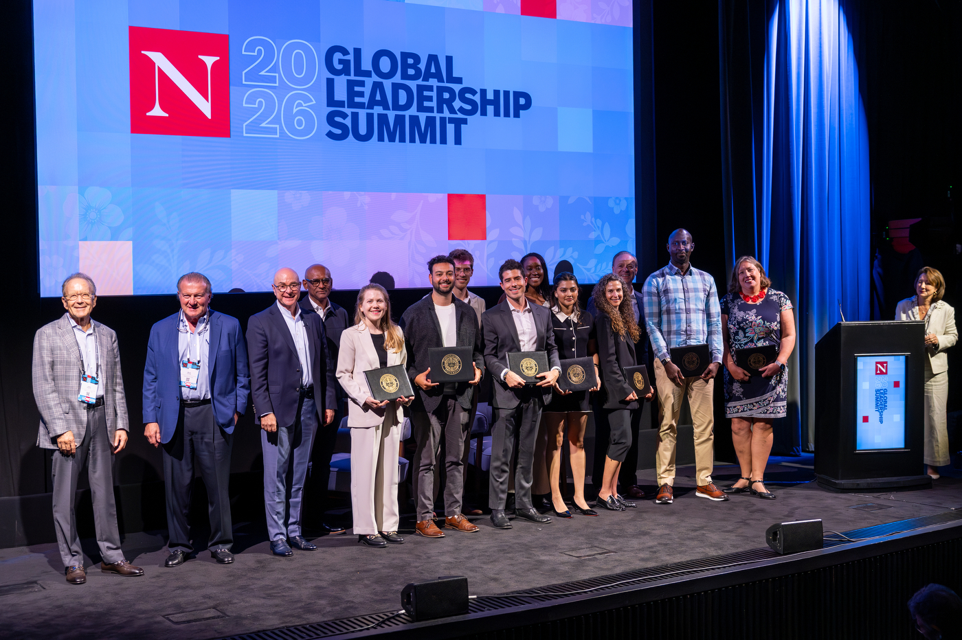 A group of people pose on stage at the Northeastern Global Leadership Summit, several holding certificate folios, with the summit logo projected on a large screen behind them.
