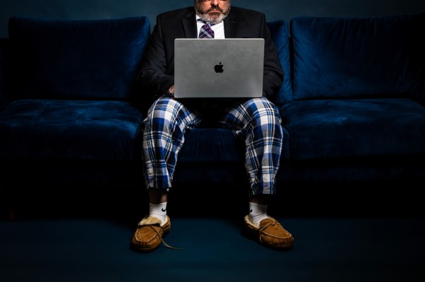 A person sitting on a couch works on a laptop, while wearing business professional clothing on top and pajama pant bottoms.