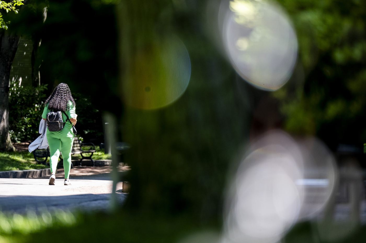 A student in a bright green outfit walks briskly along a shaded campus path, with soft bokeh light effects and green foliage in the background.