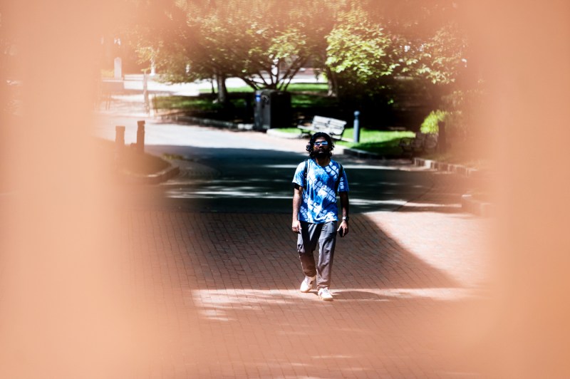 A person in a blue patterned shirt walks along a sunny brick campus path, framed by a circular vignette with trees and benches visible in the background.