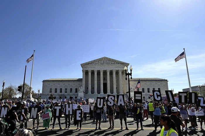 Crowd of demonstrators holding large individual letter signs spelling out messages in front of the U.S. Supreme Court building, with American flags flying on either side.