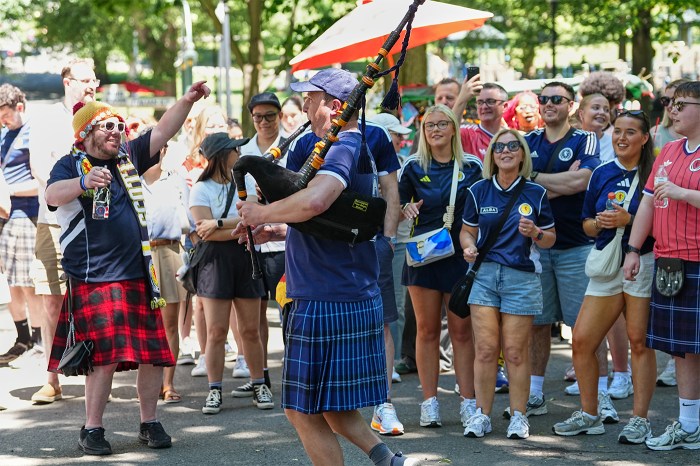 Scottish fans in kilts and blue face paint play bagpipes and celebrate among a crowd at a park in the United States.