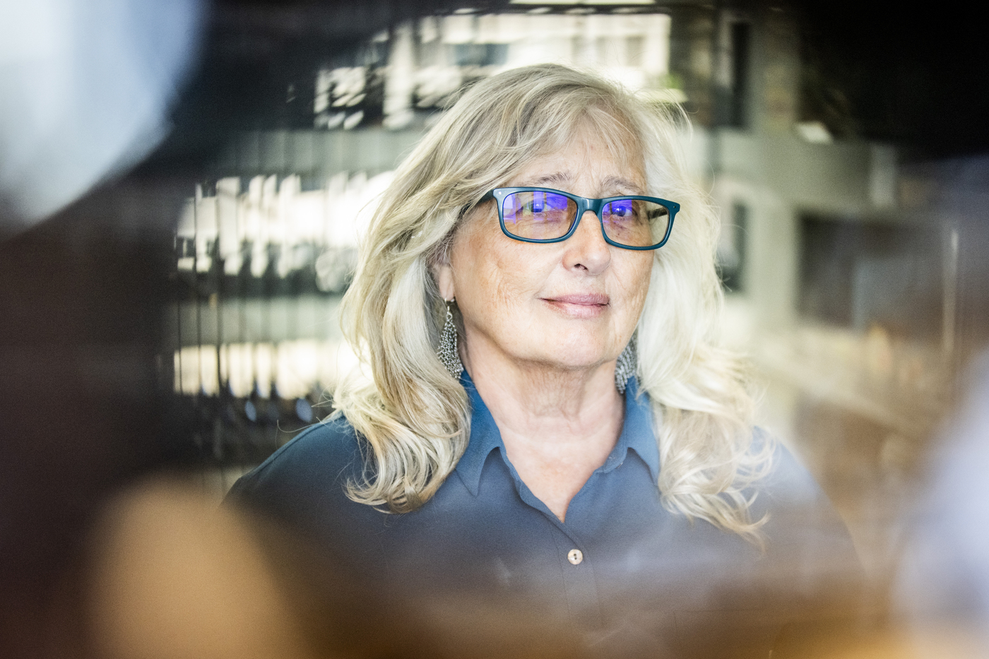 Portrait of Kathleen Kelly wearing a blue button down and green glasses in a modern building.