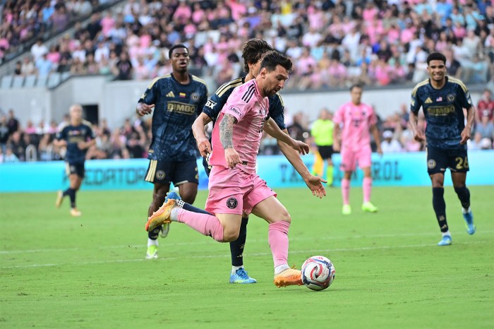 Lionel Messi wearing a pink uniform dribbles the ball during a match, with defenders closing in and a packed stadium in the background.
