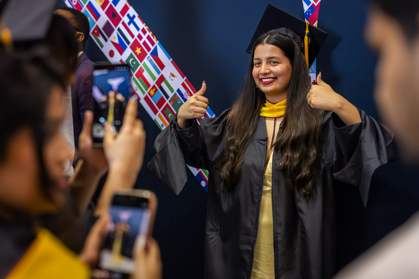 A graduate gives two thumbs up during a Northeastern commencement procession, her mortarboard decorated with international flags.