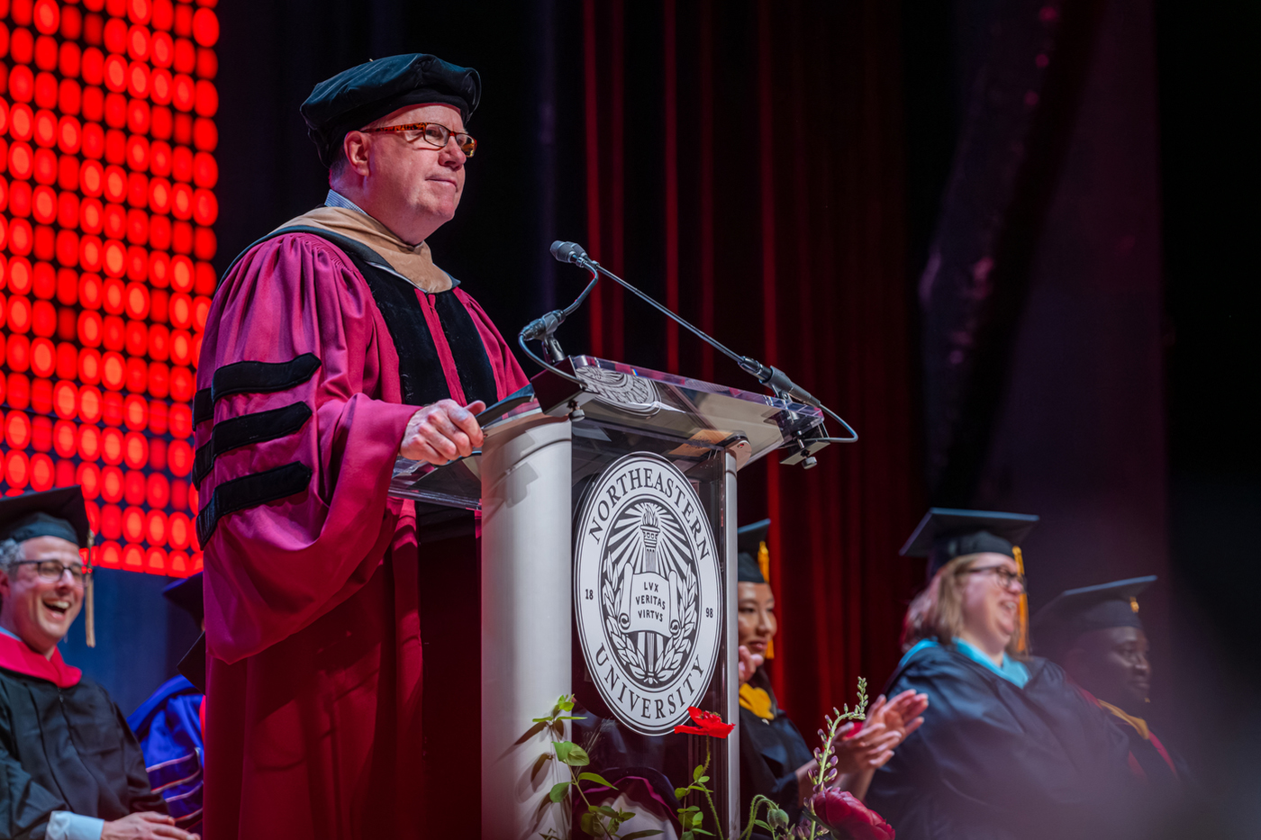 A university official in doctoral regalia speaks at the Northeastern podium during a commencement ceremony.