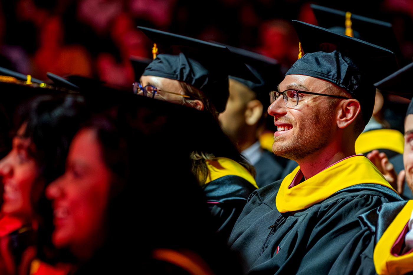 A graduate smiles during a Northeastern commencement ceremony, seated among fellow graduates in cap and gown.