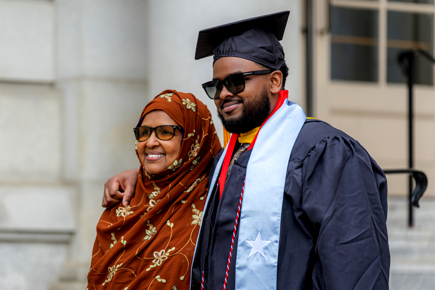 A graduate in cap and gown poses with a family member outside a commencement venue.