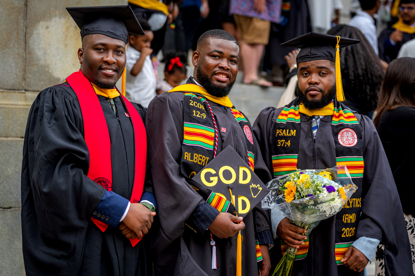 Three graduates pose together on the steps outside a commencement venue, wearing kente cloth stoles and decorated mortarboards.