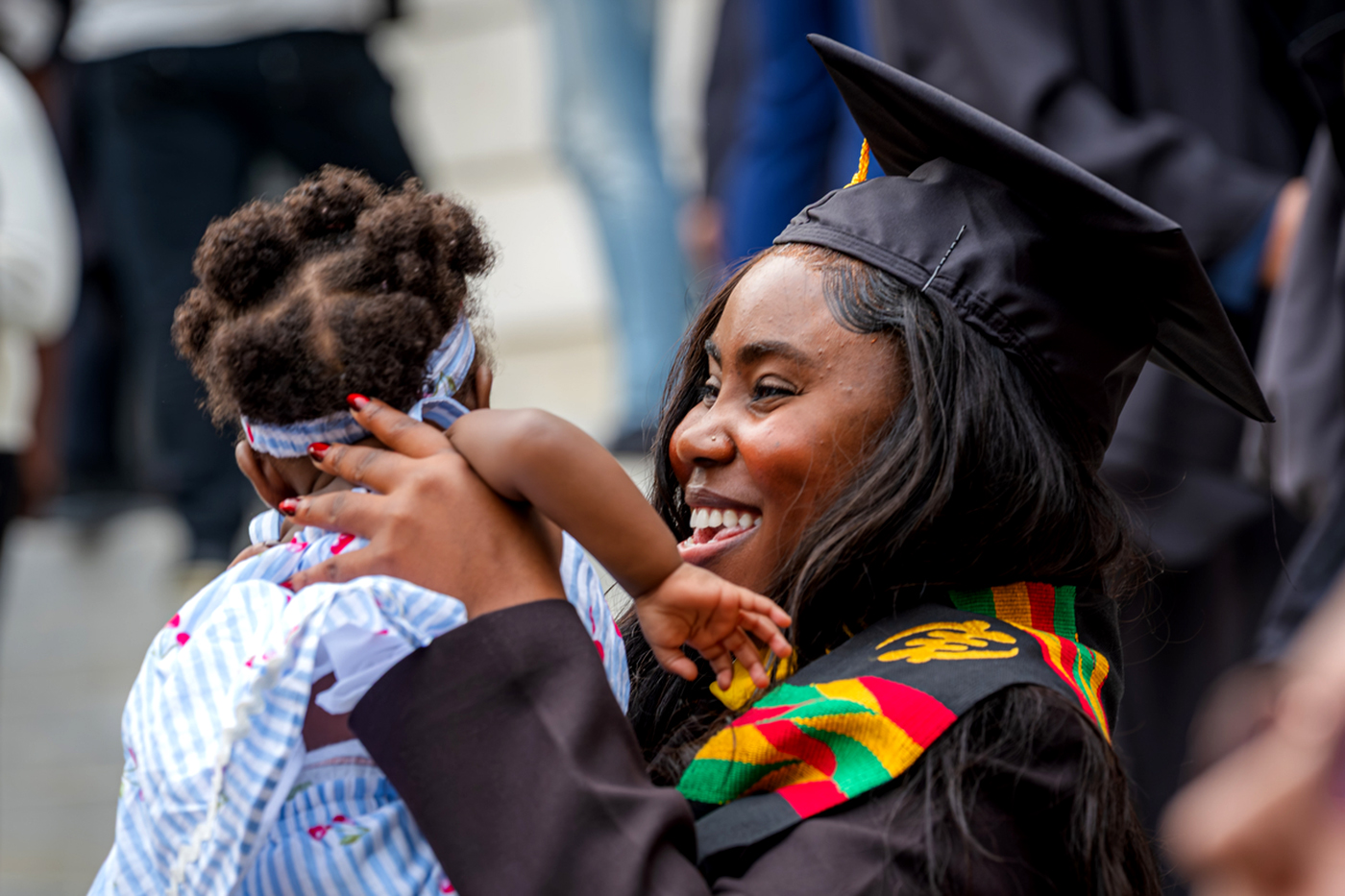 A graduate in cap and gown with a kente cloth stole embraces a child after a Northeastern commencement ceremony.