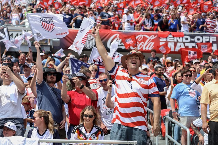 U.S. soccer fans cheering in the stands, one waving a USMNT flag, at a packed stadium during a World Cup send-off match.