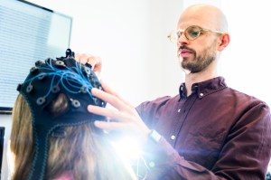 Claude responded: A researcher adjusts an EEG cap fitted on a study participant's head, with a computer monitor visible in the background.A researcher adjusts an EEG cap fitted on a study participant's head, with a computer monitor visible in the background.