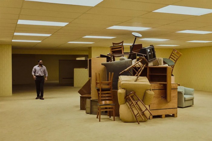 A man stands alone in a large, nearly empty room facing a towering pile of furniture — chairs, sofas, shelves, and a television — stacked in the center under f…A man stands alone in a large, nearly empty room facing a towering pile of furniture — chairs, sofas, shelves, and a television — stacked in the center under fluorescent lights.