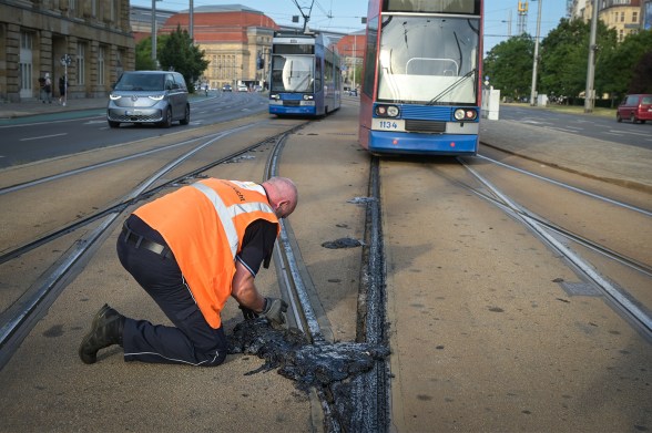 A worker in an orange safety vest examines heat-damaged tram tracks in Leipzig, where melted bitumen buckled under extreme temperatures as trams pass in the background.