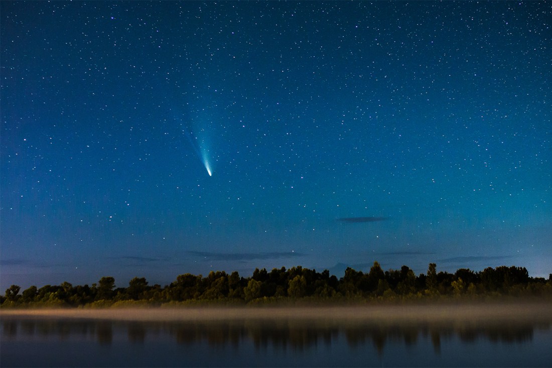 A bright-tailed comet streaks across a starry night sky above a fog-covered river and treeline.