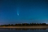 A bright-tailed comet streaks across a starry night sky above a fog-covered river and treeline.