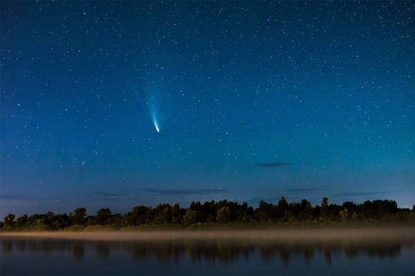 A bright-tailed comet streaks across a starry night sky above a fog-covered river and treeline.