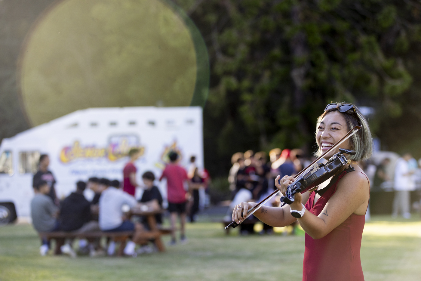 A violinist performs outdoors at an event, with people gathered near a food truck in the background.