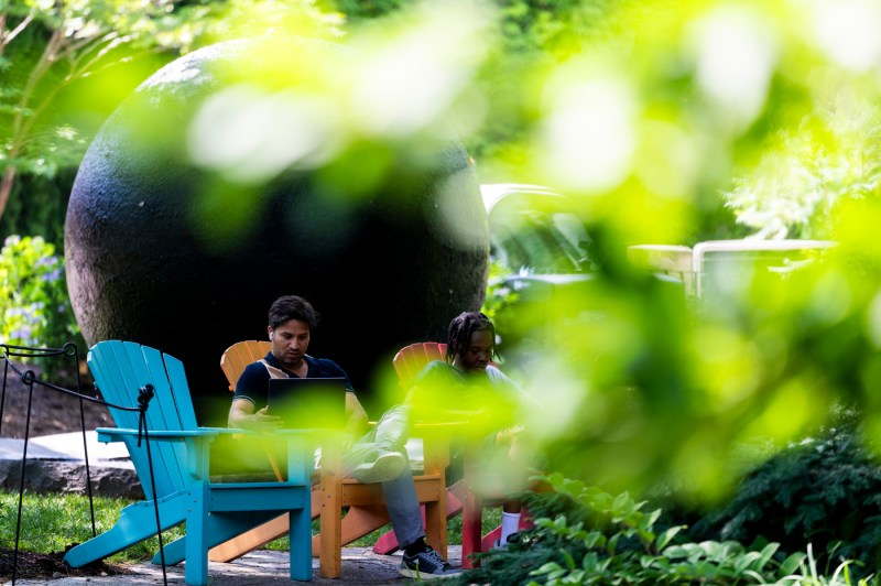 Students sit at an outdoor table under leafy trees, studying near a large dark stone sphere sculpture beside a koi pond.