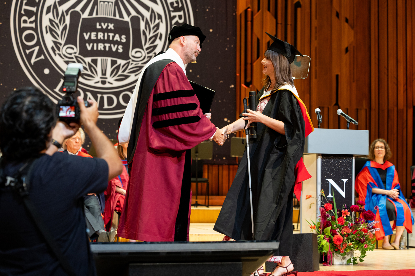 A wider view of Alexandra shaking hands with the university official on stage as a photographer documents the moment and another graduate waits at a podium.