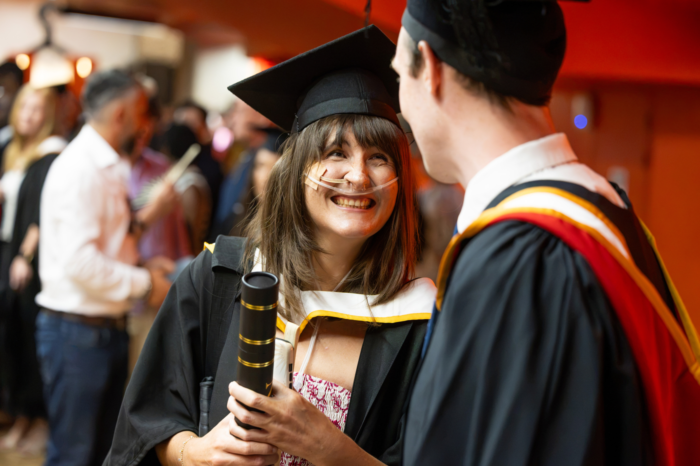 Alexandra, holding her diploma case, smiles up at another graduate as they talk at an indoor reception.
