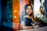 Shashi Murthy wearing a black top, seen inside of a building with warm lighting through the glass of a window.