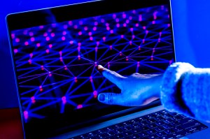 A hand reaches toward a laptop screen displaying a glowing network of connected nodes, representing quantum computing.
