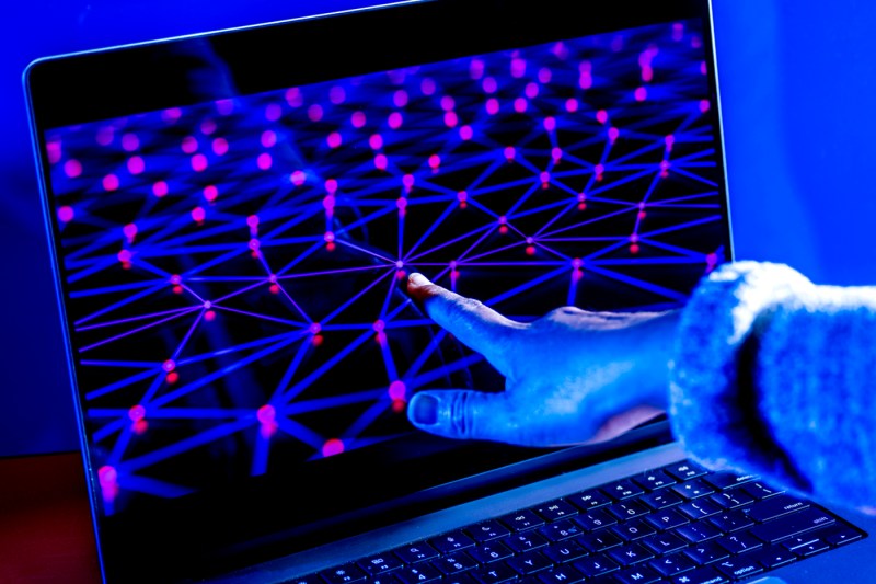 A hand reaches toward a laptop screen displaying a glowing network of connected purple and red points, lit in blue.
