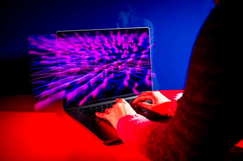 A person types on a laptop as an illuminated 3D data visualization projects above the screen, lit in red and purple.

