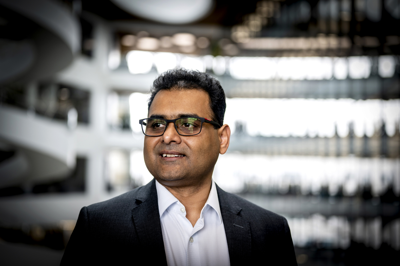 Ayan Paul, research associate professor at Northeastern University, smiling in an indoor space with a blurred staircase and shelving behind him.