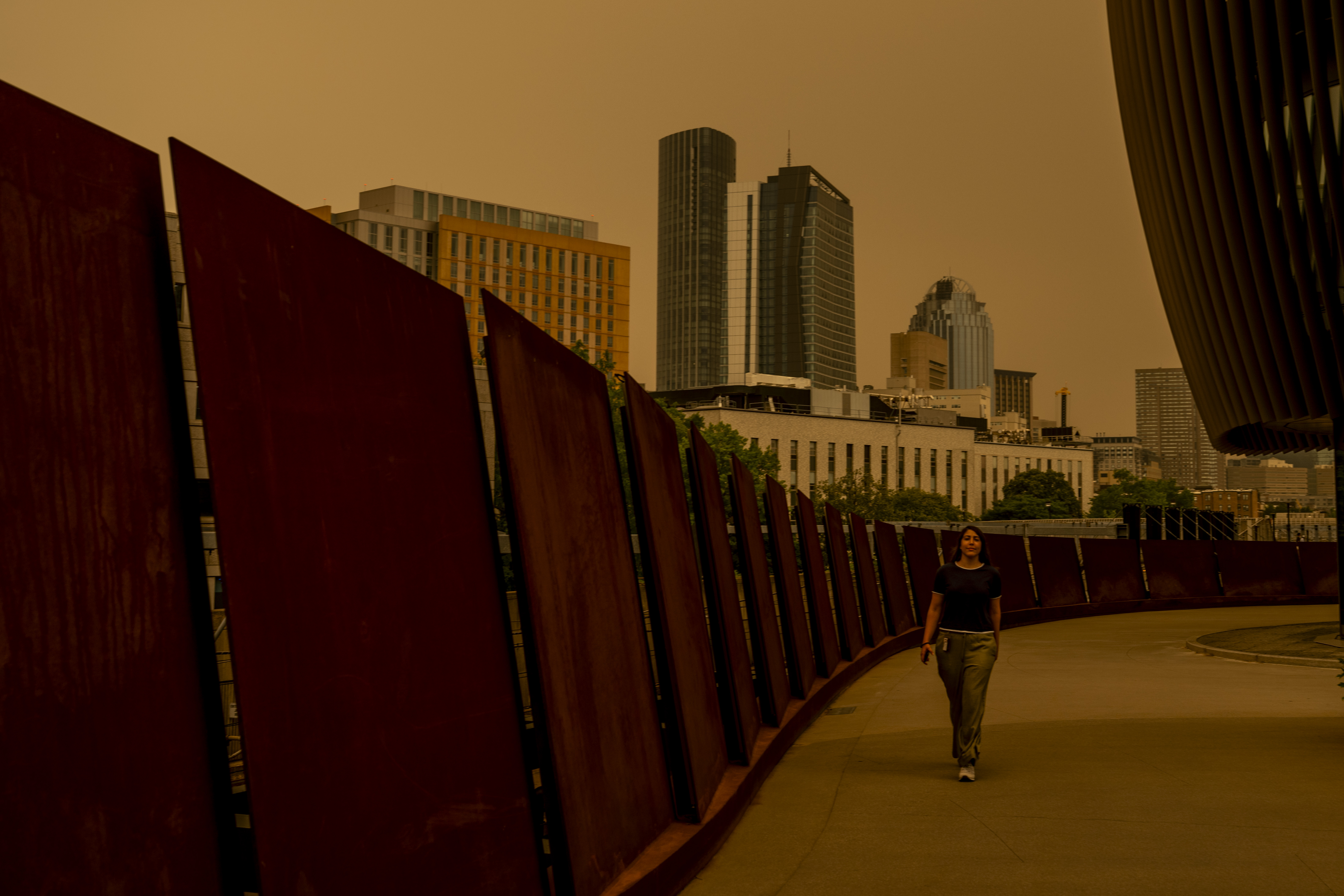 An orange, smoke-filled sky hangs over the Boston skyline as a person walks by.
