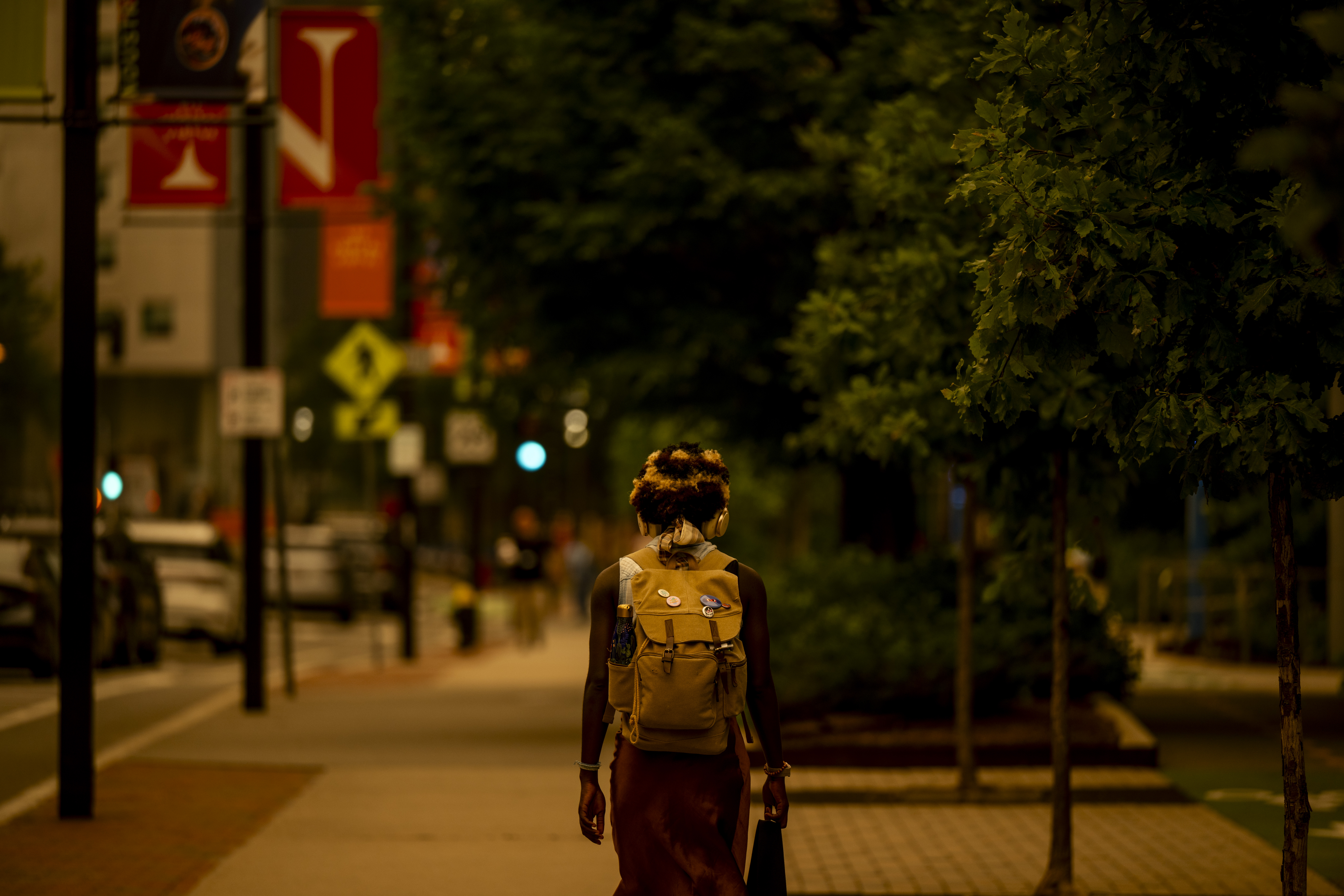 A student wearing a backpack walks down a city sidewalk under a hazy orange sky at dusk.


