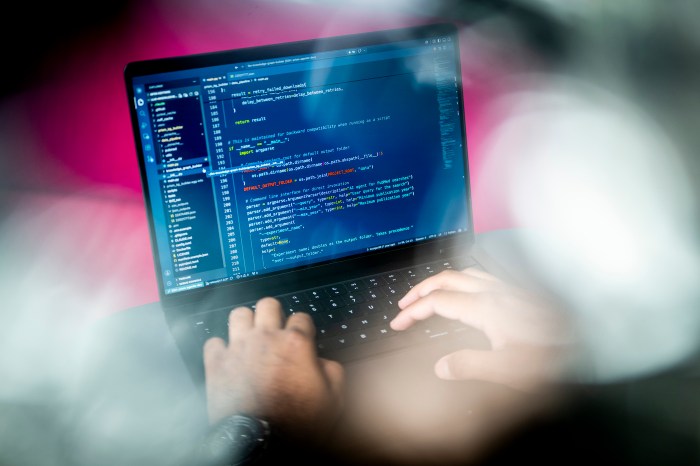 A person's hands type on a laptop displaying Python code, with a soft-focus foreground of pink and white light.