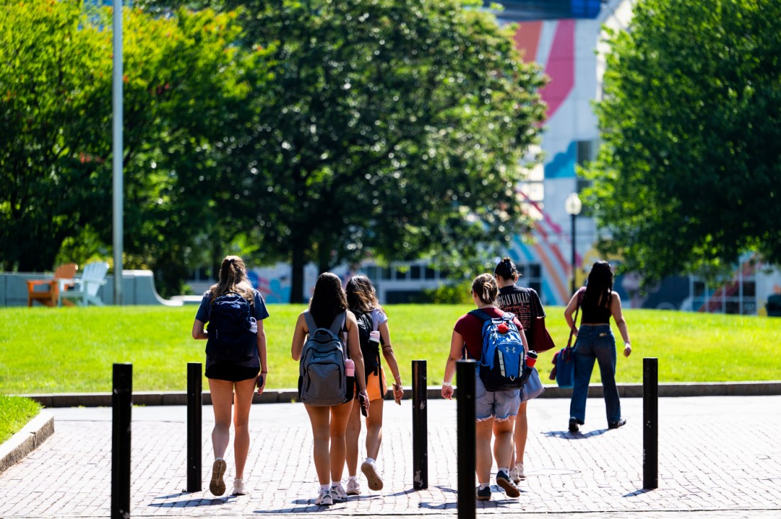 A group of students, some carrying backpacks, walks across an outdoor plaza with trees and a colorful mural in the background.

