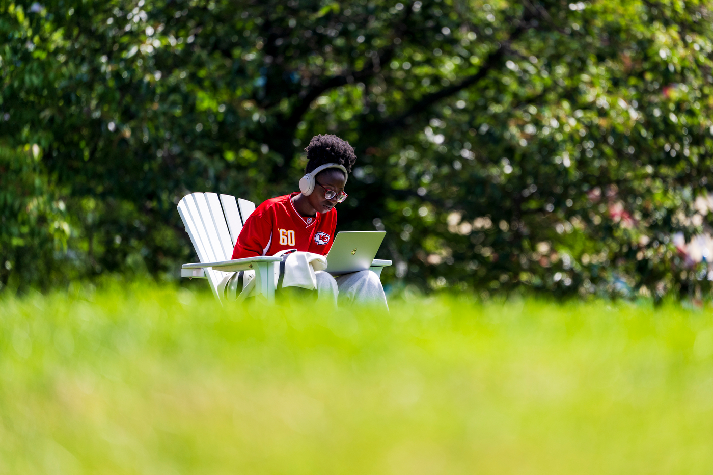 A student using a laptop sits in an Adirondack chair in a grassy field.