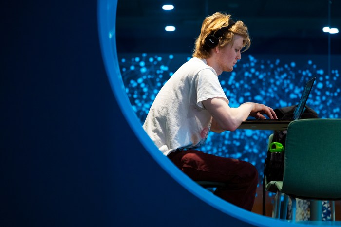  student wearing headphones works on a laptop while seated in a circular window opening, with a wall of illuminated blue particle-like lights visible behind him.