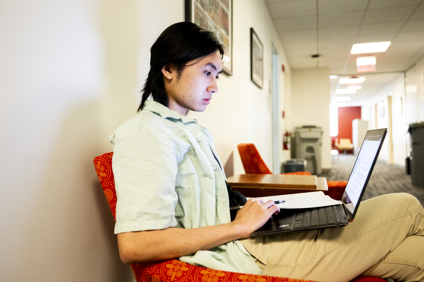 A student sits in a hallway with a laptop and notebook, writing while working.